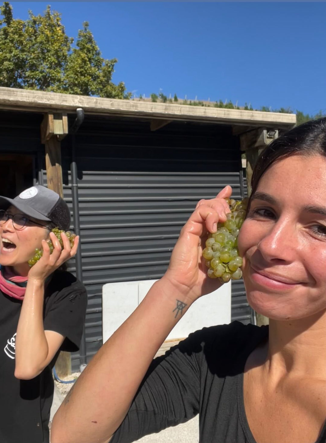 Two women outdoors in front of a dark garage door, each holding a cluster of green grapes. One woman in the foreground is smiling at the camera, and another woman is partially visible on the left.